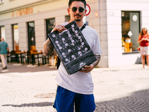 Ein tätowierter Mann hält eine Tasche in der Hand und blickt direkt in die Kamera.