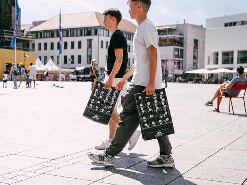 Zwei Jungen gehen auf einem Bürgersteig, umgeben von Bäumen und einem klaren Himmel.