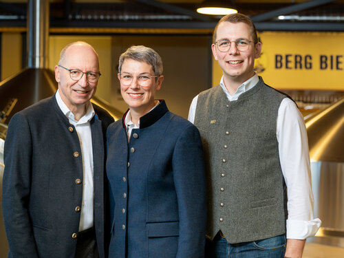 Eine Frau und zwei Männer in traditioneller bayerischer Tracht stehen zusammen und lächeln freundlich in die Kamera.
