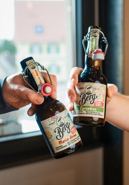 Zwei Personen halten Bierflaschen vor einem Fenster und genießen gemeinsam den Moment.